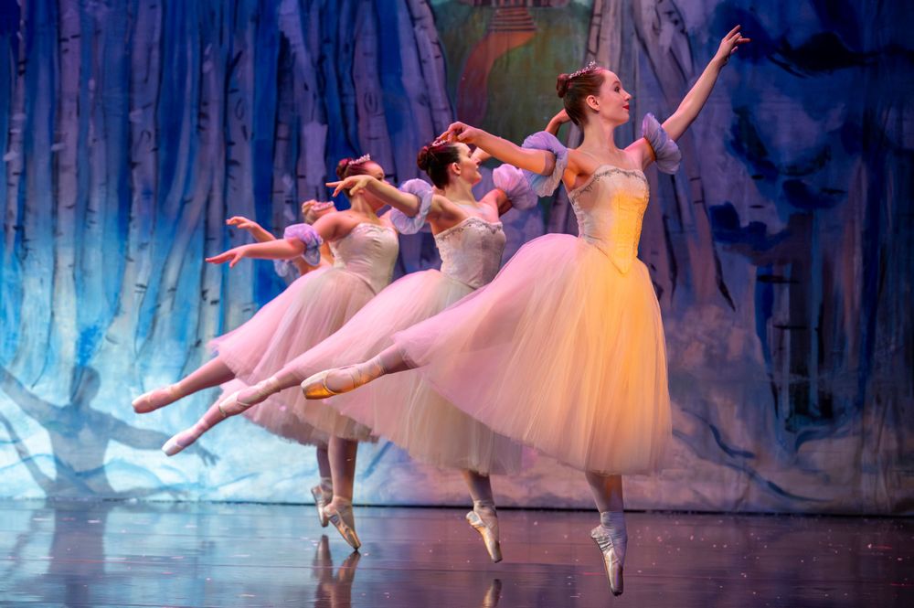 Ballet dancers in white tutus performing The Nutcracker on stage with snowy forest backdrop.
