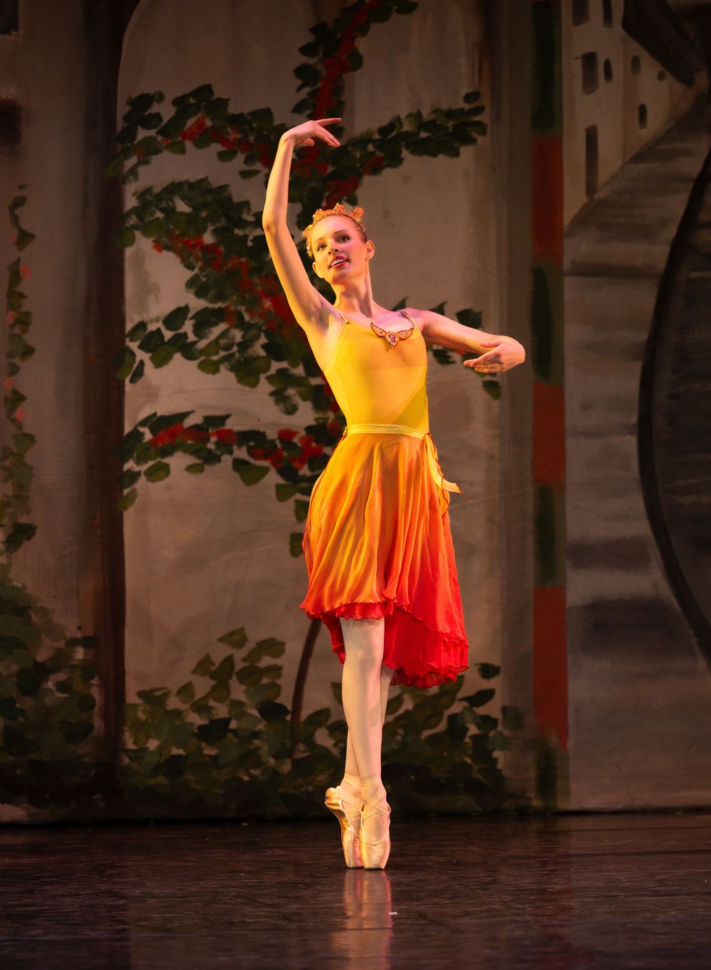 Ballerina in yellow and orange gradient costume performs en pointe on stage with floral backdrop.