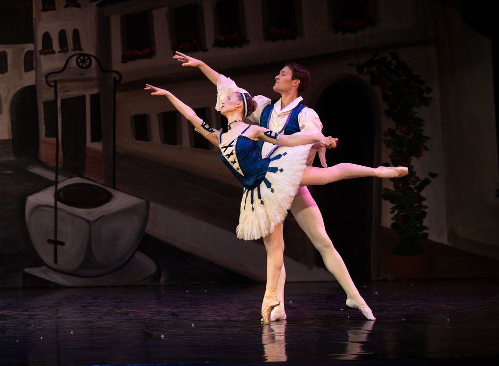 Two ballet dancers perform a graceful pas de deux on stage in costume before a painted backdrop.