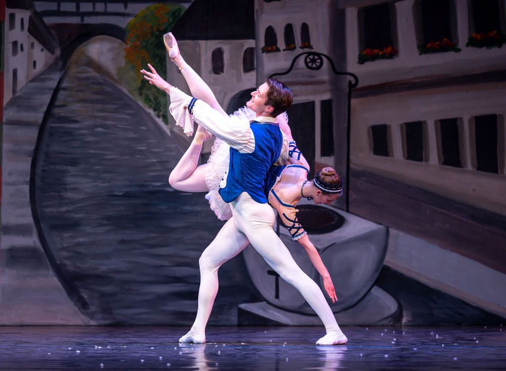 Two ballet dancers perform a graceful lift on stage with a painted European cityscape backdrop.