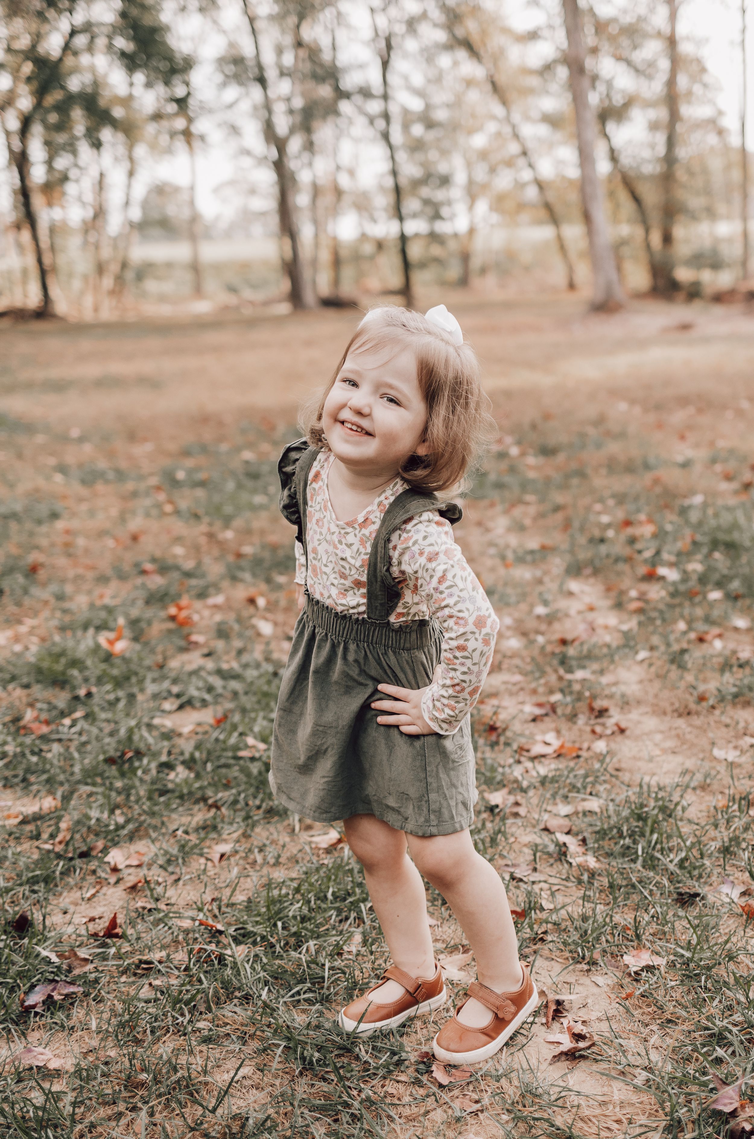 A toddler wearing a green romper and lace top plays joyfully in a park during autumn as fallen leaves scatter the ground.