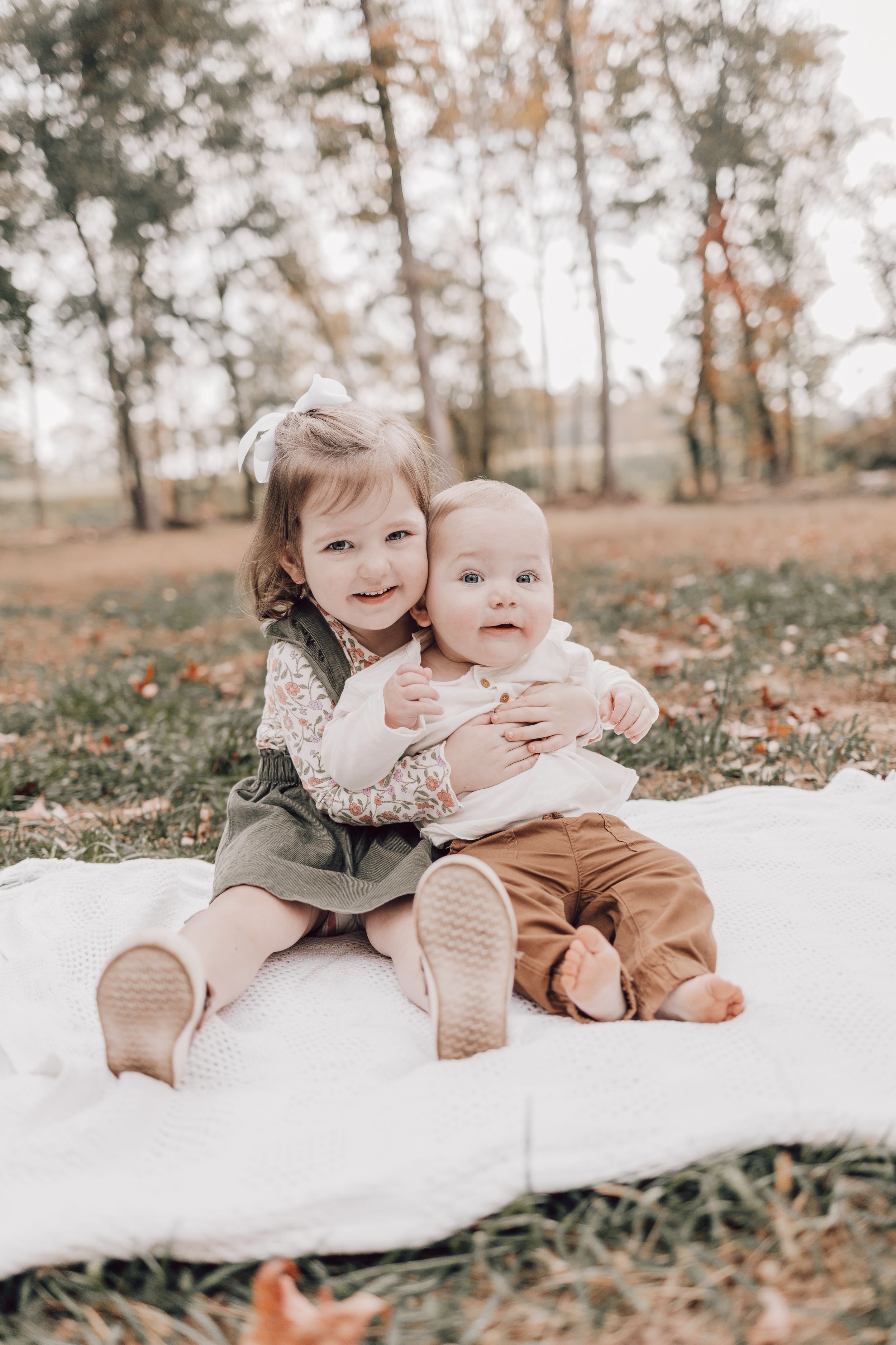 Two children sitting together on white blanket surrounded by fallen autumn leaves.