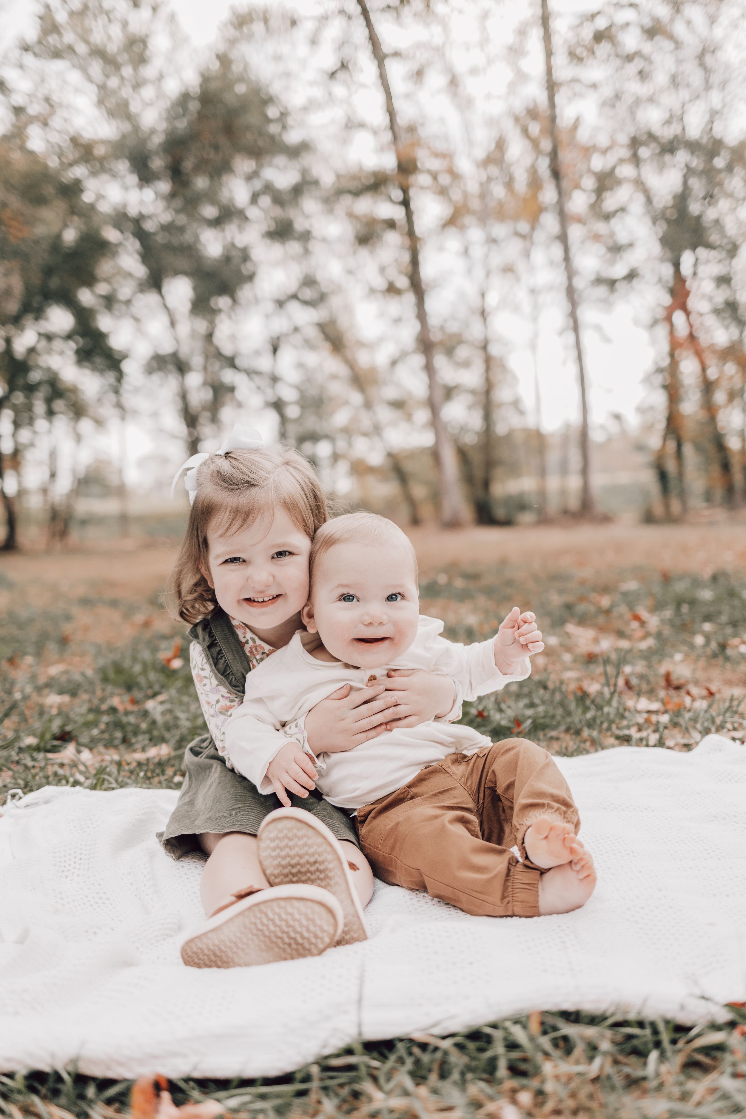 Two children sitting together on a white blanket outdoors during autumn with fallen leaves and trees in background.