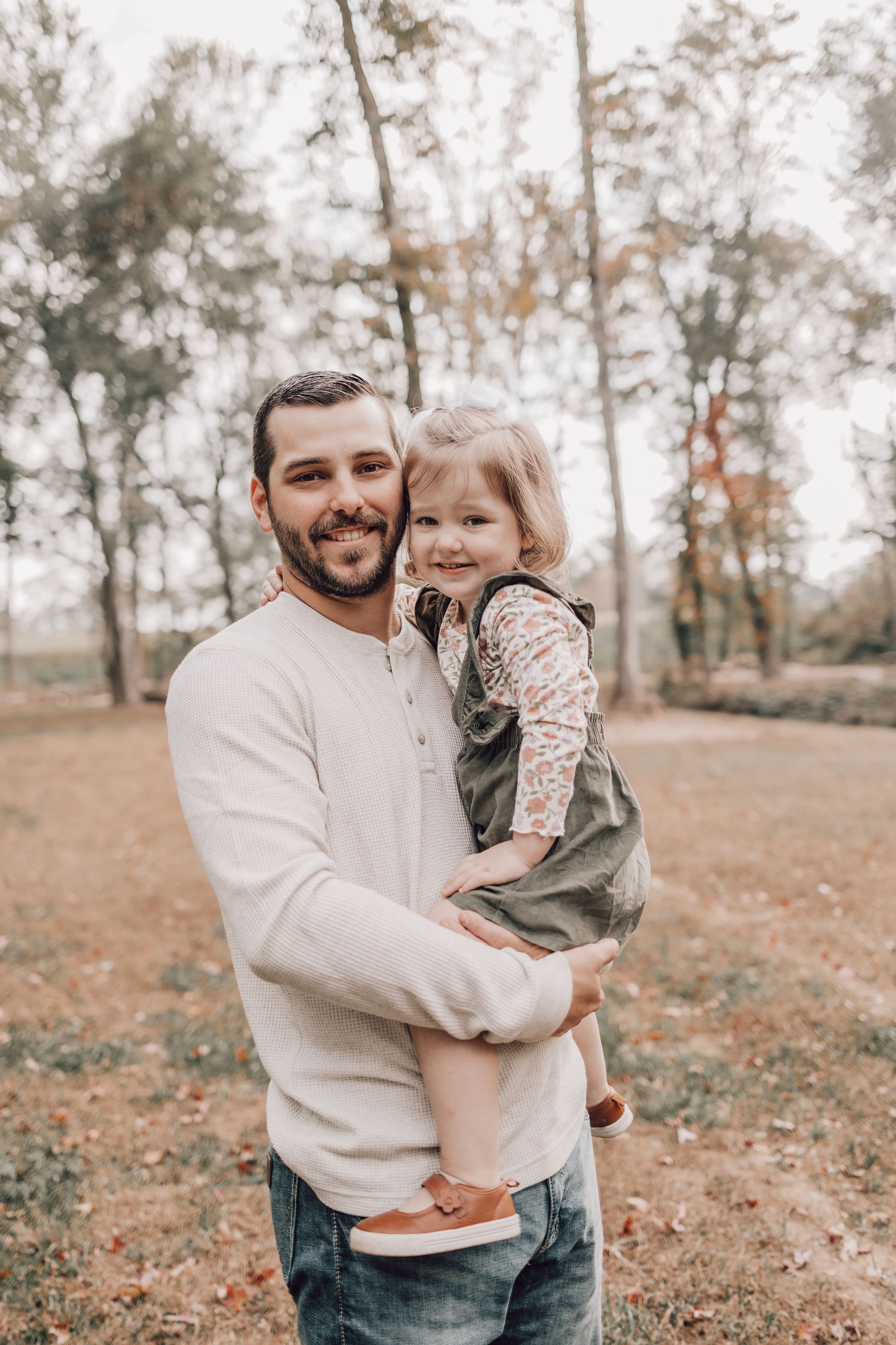 A heartwarming outdoor family portrait captured in an autumn setting with trees and fallen leaves in the background.