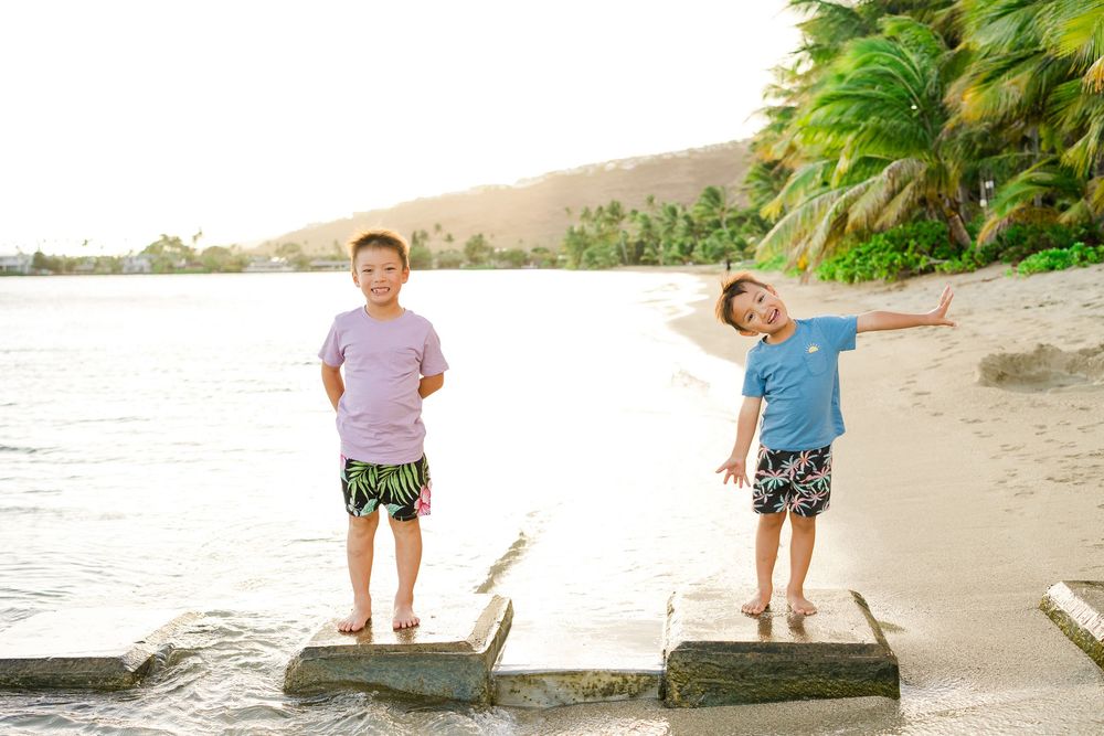 Two children stand on logs at a tropical beach during sunset with arms outstretched.