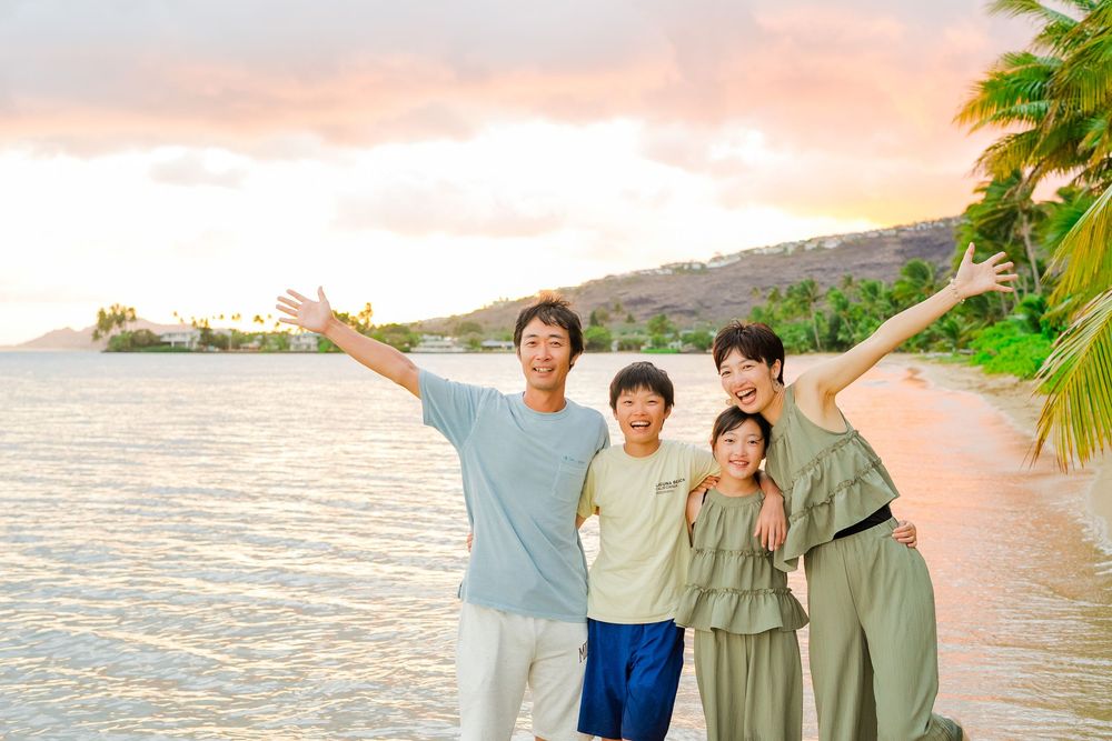 A group of people pose with arms raised on a tropical beach at sunset with palm trees and ocean in the background.