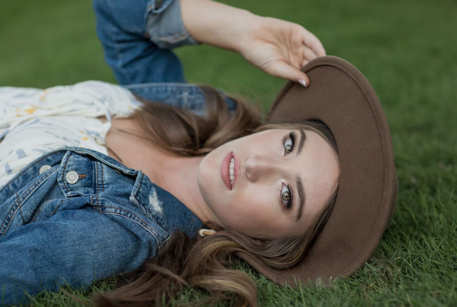 Young woman in jeans and floral top lying on grass, holding a brown cowboy hat, smiling at camera.