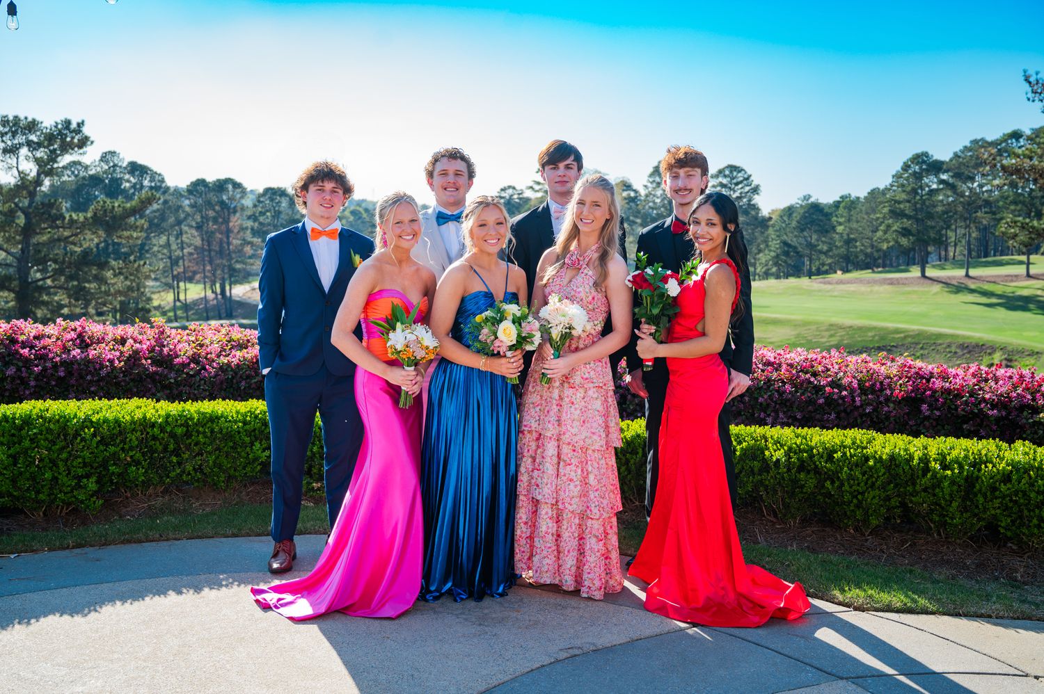 A formal group poses together in elegant evening attire against a backdrop of pink flowers and a golf course on a sunny day.