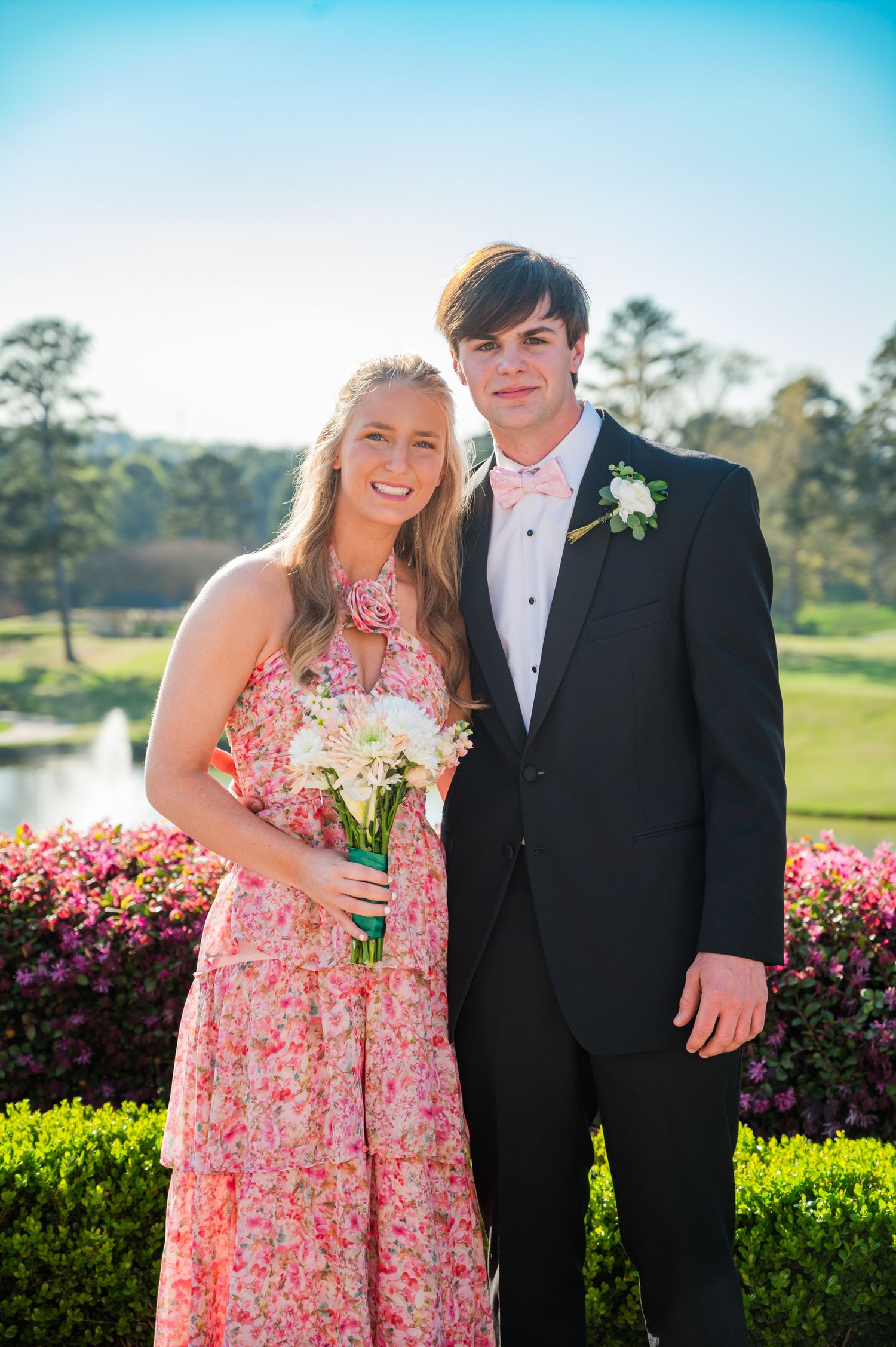 A couple dressed in formal attire pose together by blooming pink flowers on a sunny spring day.