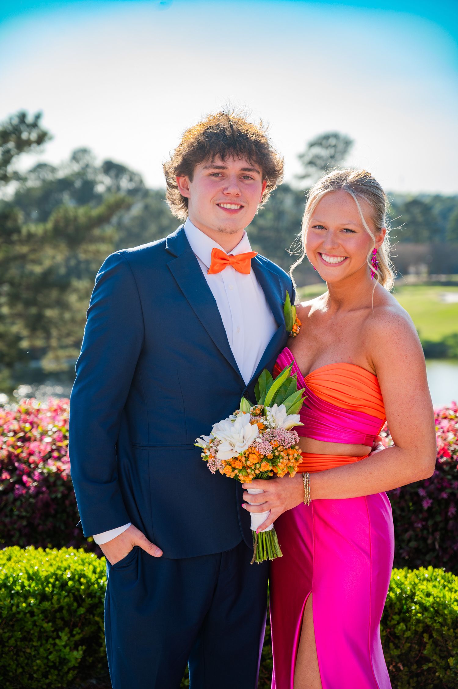 A couple in formal attire poses together at an outdoor event, with the woman in a bright pink dress holding a bouquet.