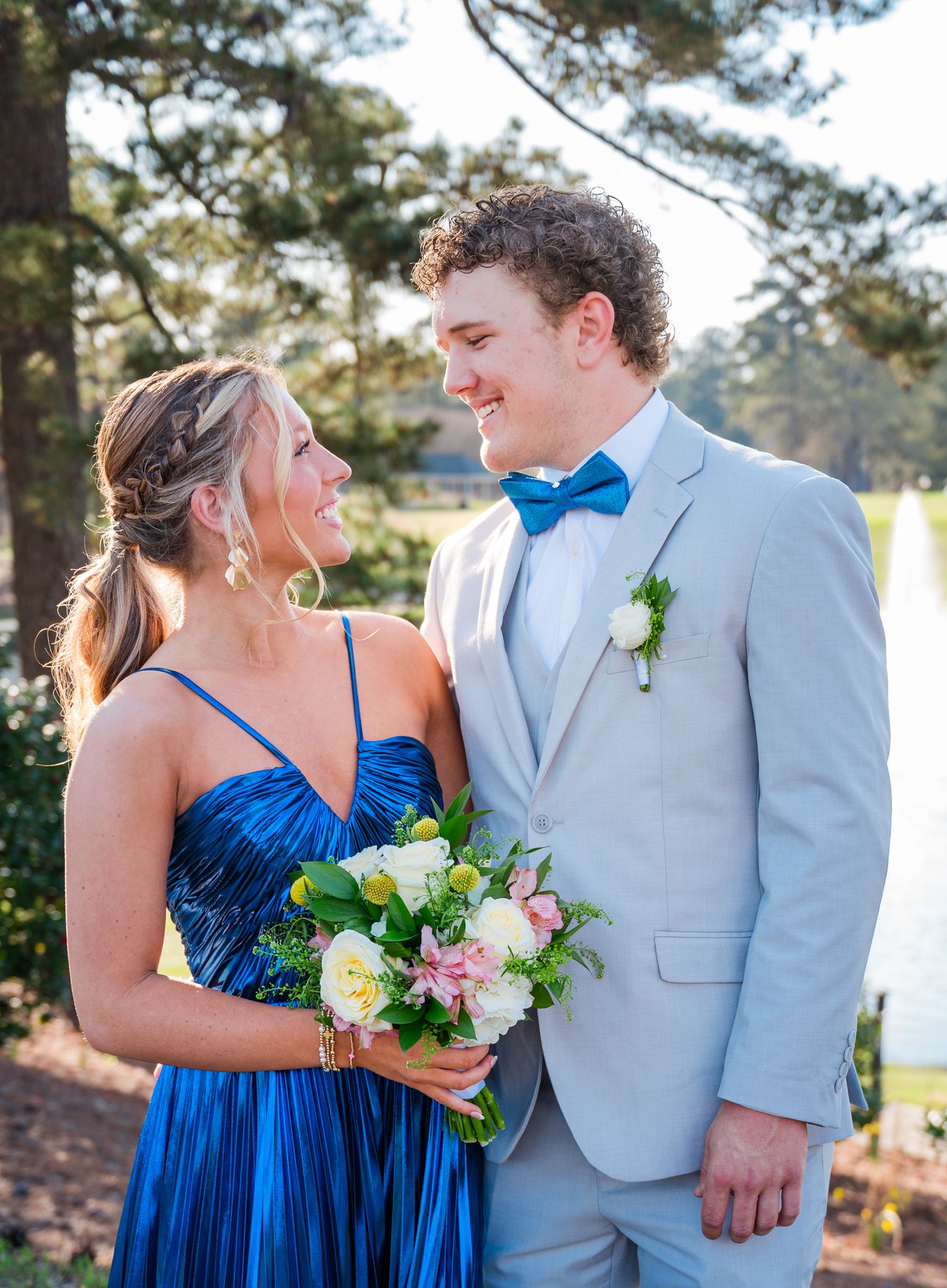 A couple dressed in a royal blue dress and light gray suit pose together outdoors with a floral bouquet.