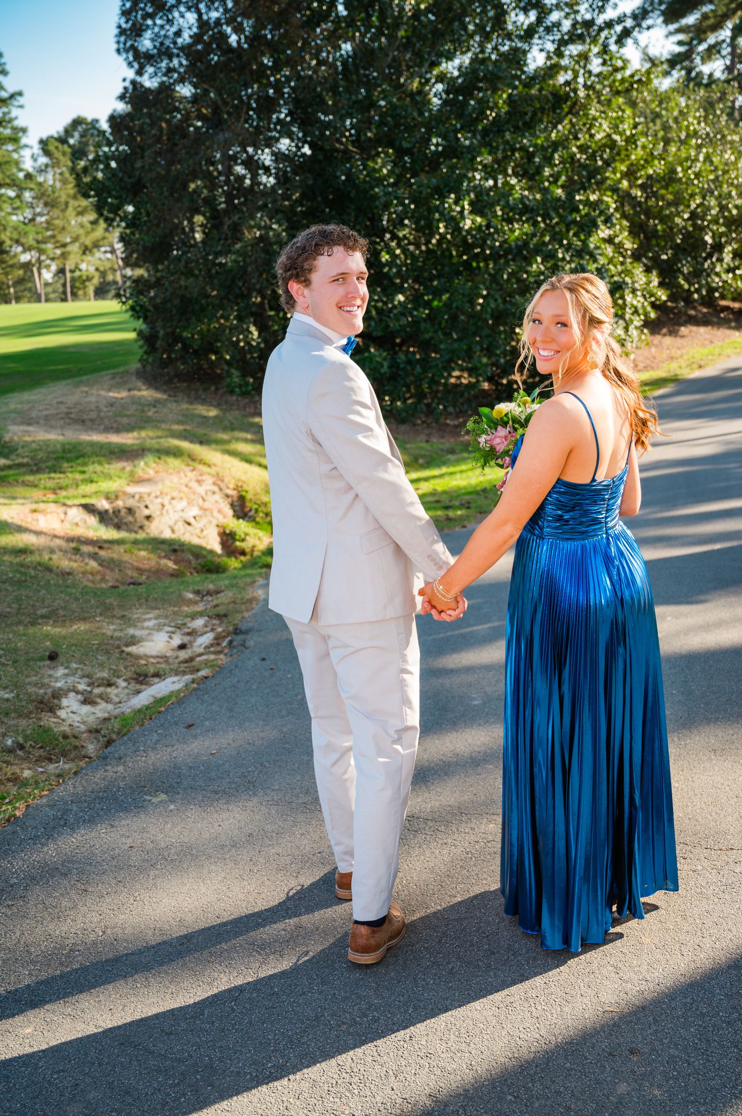 A couple holding hands in formal attire walks on a path, with a blue dress and light gray suit against a sunny outdoor setting.