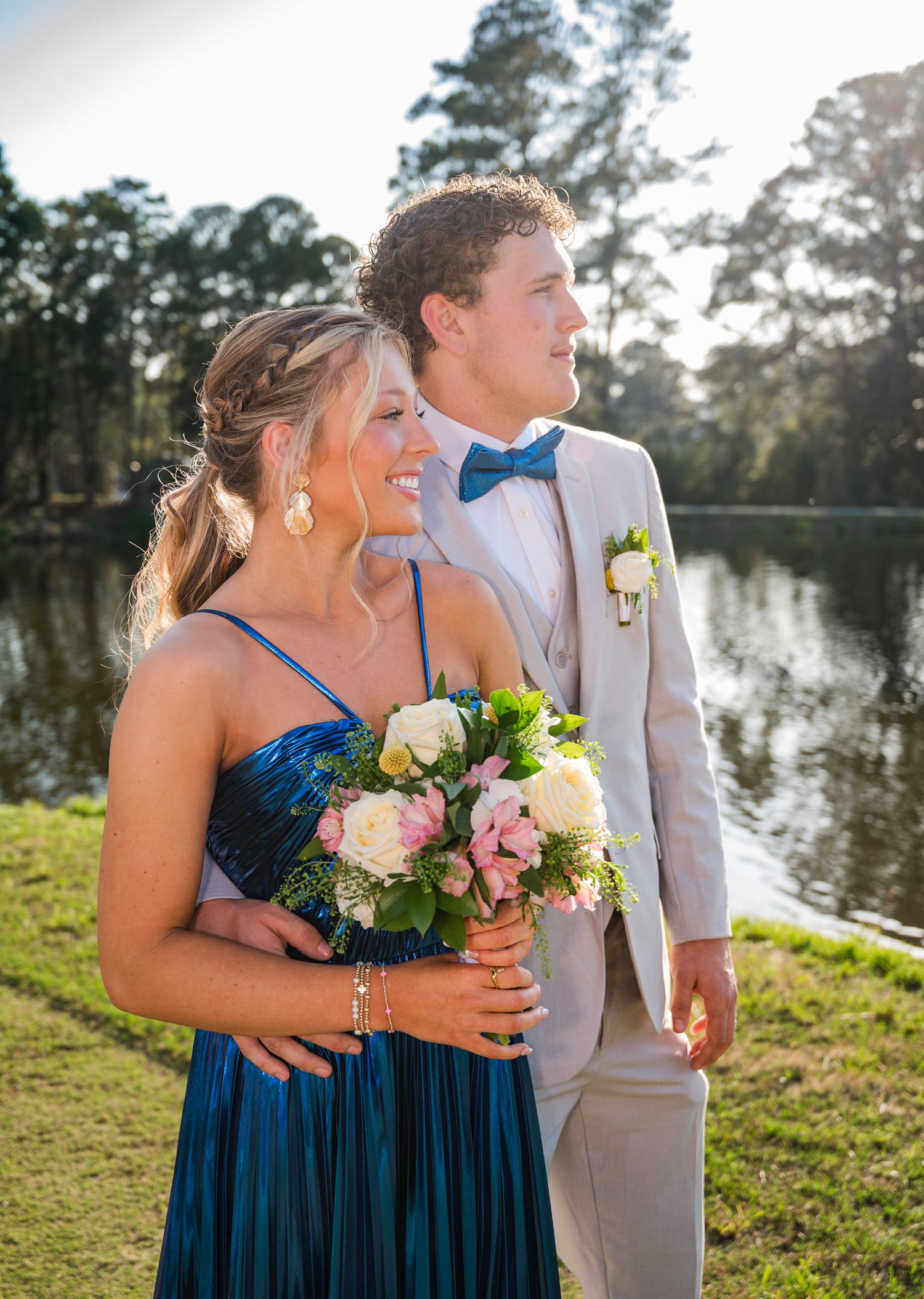 A couple in formal attire poses by a lake, with the bridesmaid in navy blue holding pink and white flowers.