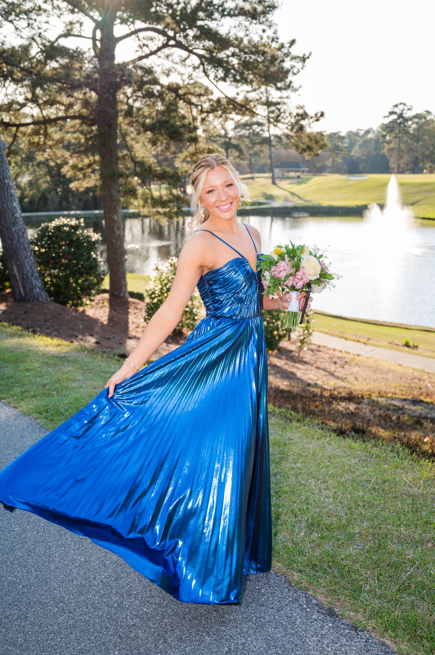 A person in a flowing metallic blue gown twirls on a path near a lake at sunset with pine trees in the background.