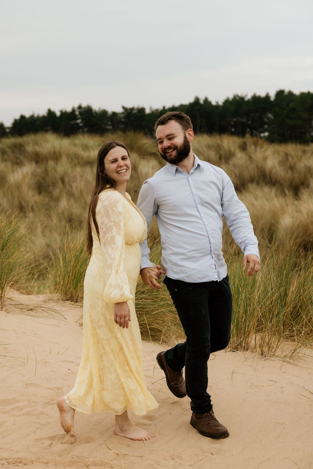 Couple walking hand in hand on sandy dunes, woman in yellow lace dress, man in light blue shirt, green trees behind.