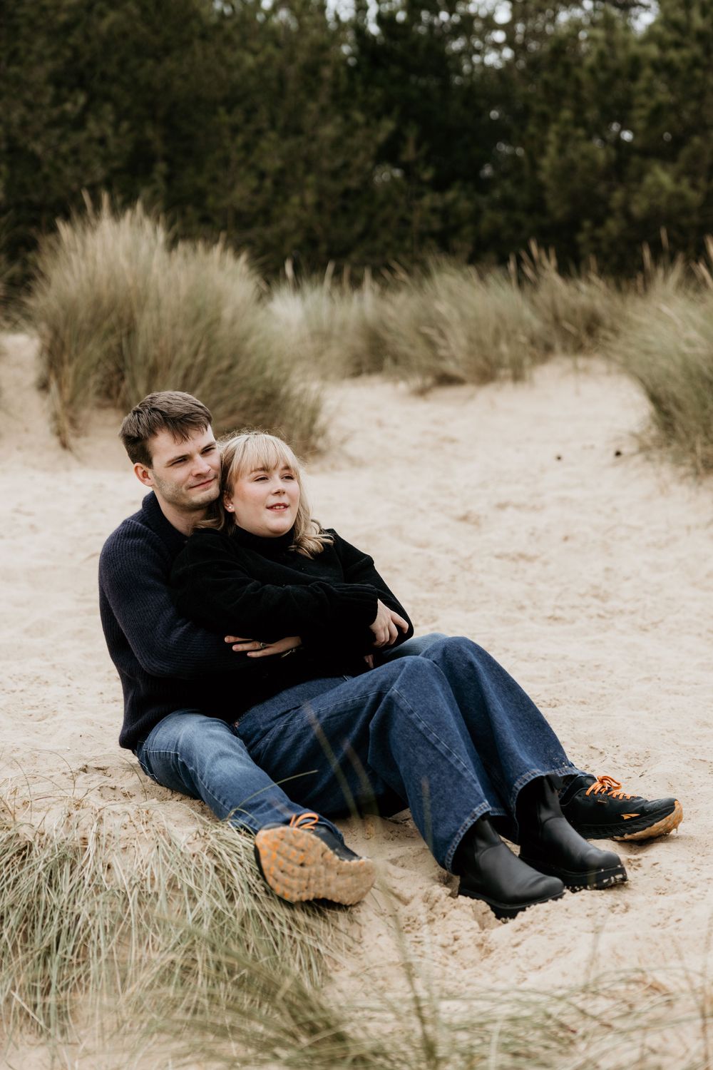 Couple sitting together on sandy beach dunes, wearing dark sweaters and jeans, embracing warmly with green grass in background.
