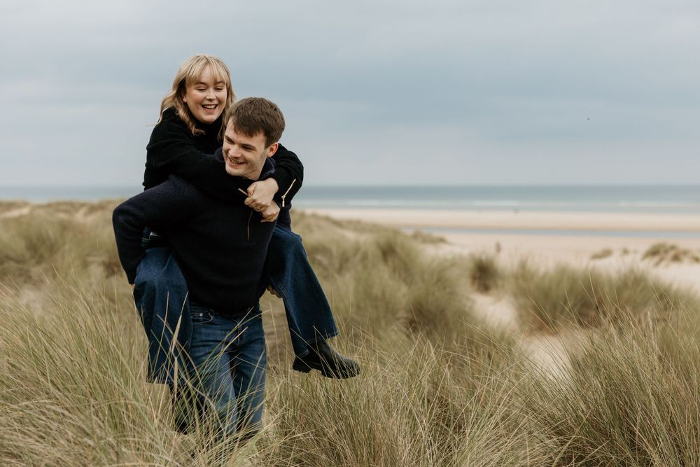 A couple laughing on sandy beach dunes, man giving woman a piggyback ride with ocean in background.
