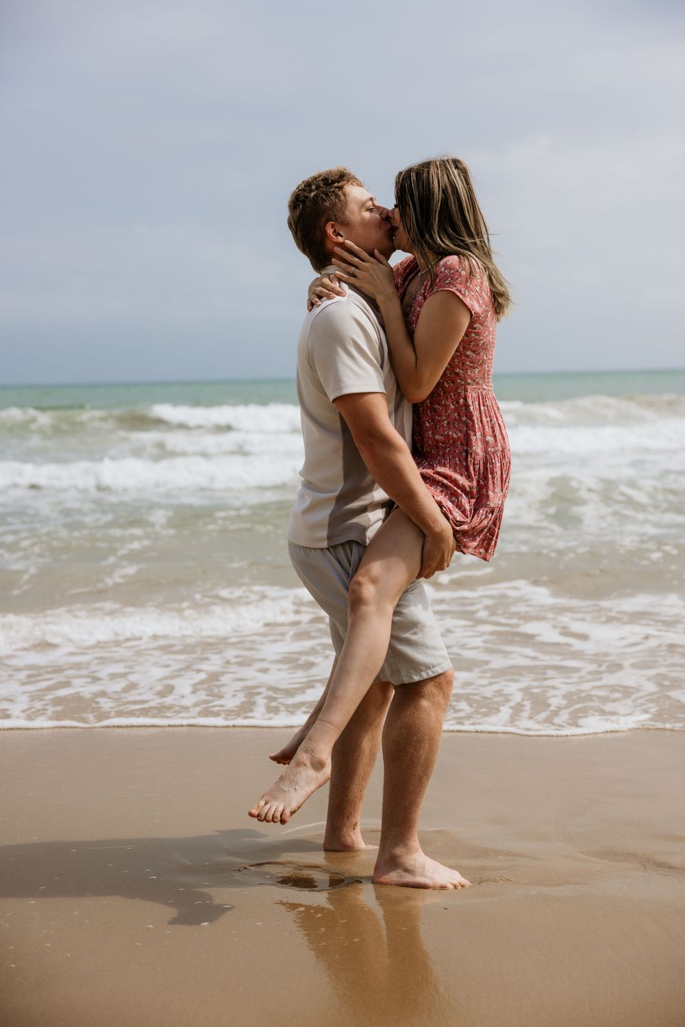 A couple shares a romantic kiss on a sandy beach, with ocean waves in the background on a cloudy day.