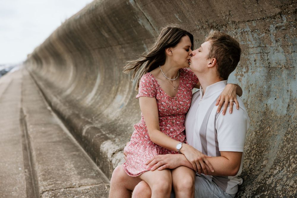 A couple sharing a romantic moment, nearly kissing while sitting against a concrete wall outdoors.