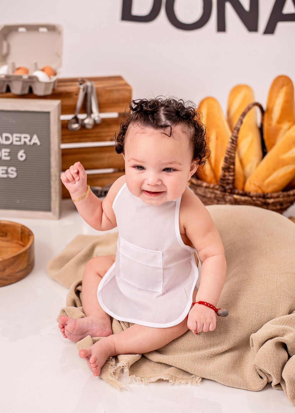 Happy baby in white bib sits on burlap near bread basket and eggs in a charming bakery-themed photo setup.