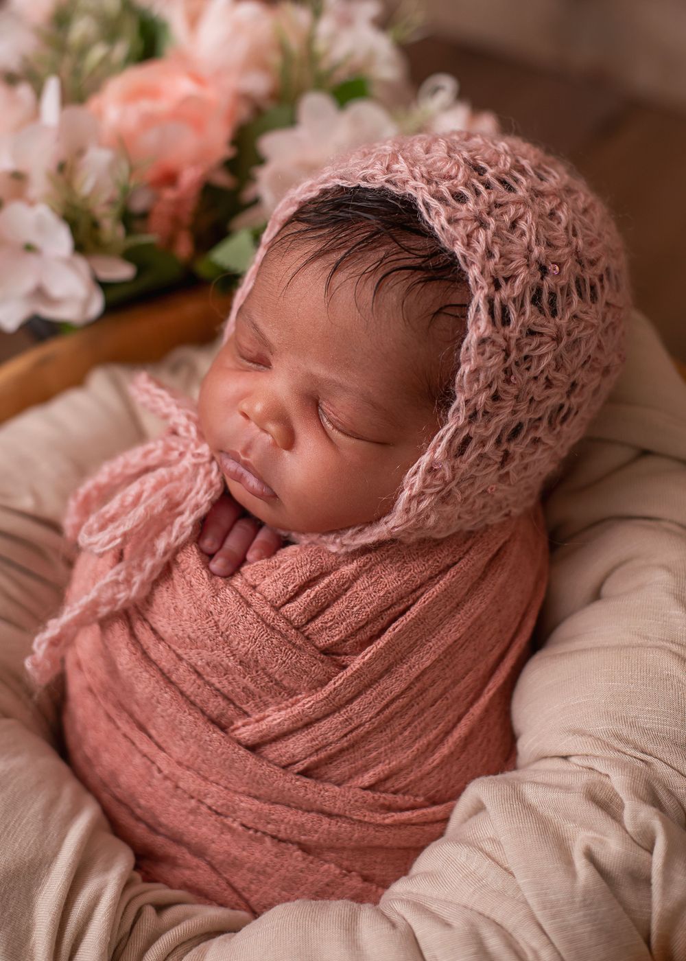 Sleeping newborn baby swaddled in dusty rose wrap wearing a pink knit bonnet, nestled in a basket with flowers.