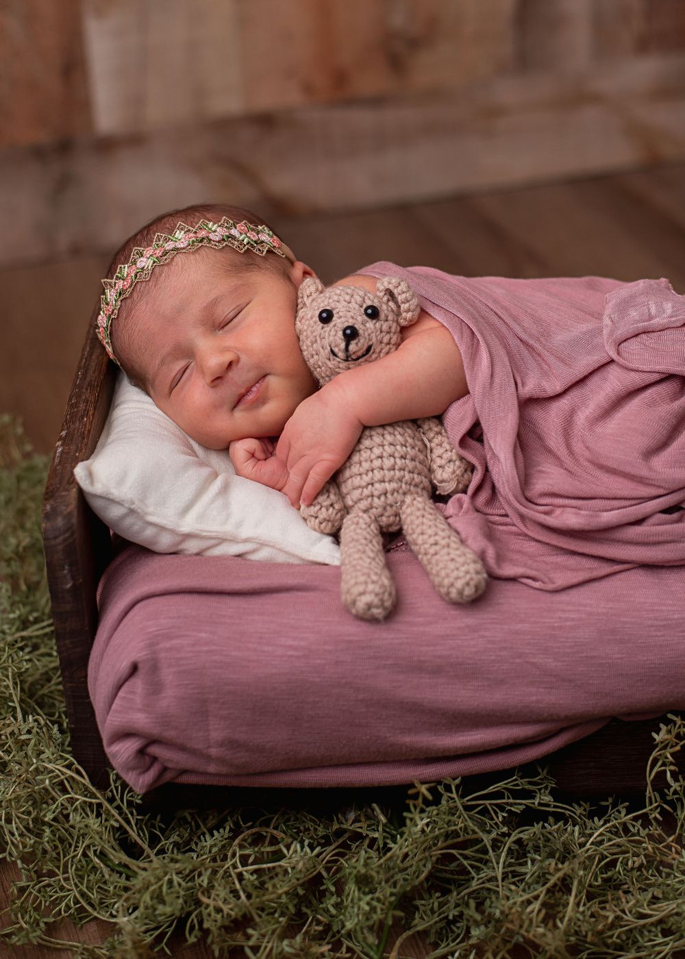 Sleeping newborn baby in pink wrap holding crochet bear toy in rustic wooden bed with white pillow.