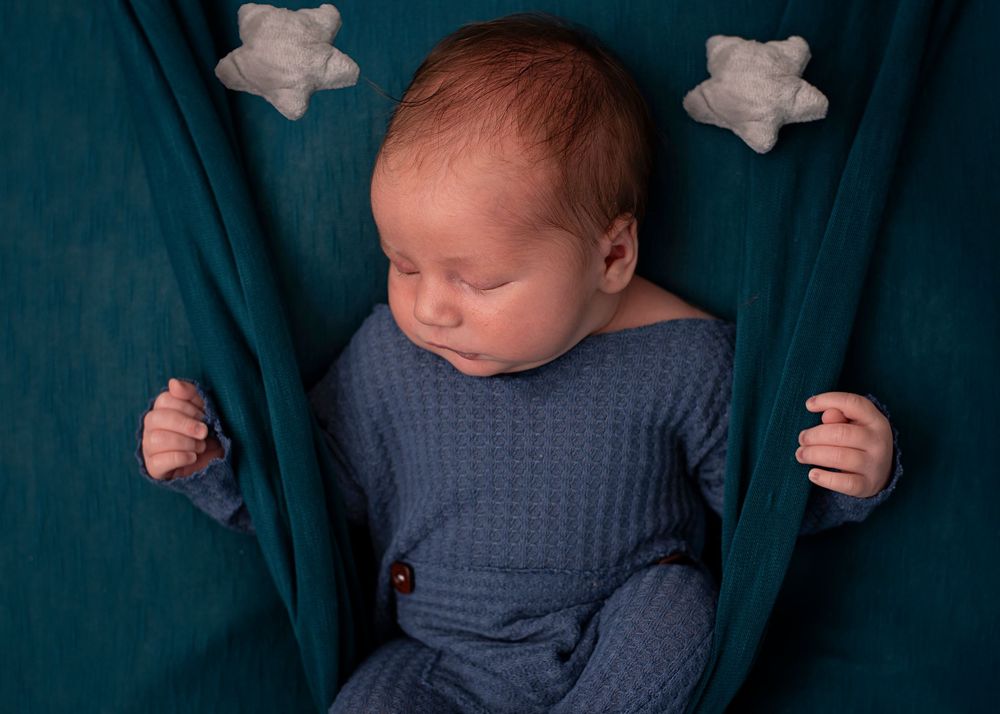 Sleeping newborn baby in blue knit outfit on teal fabric backdrop with white star props.