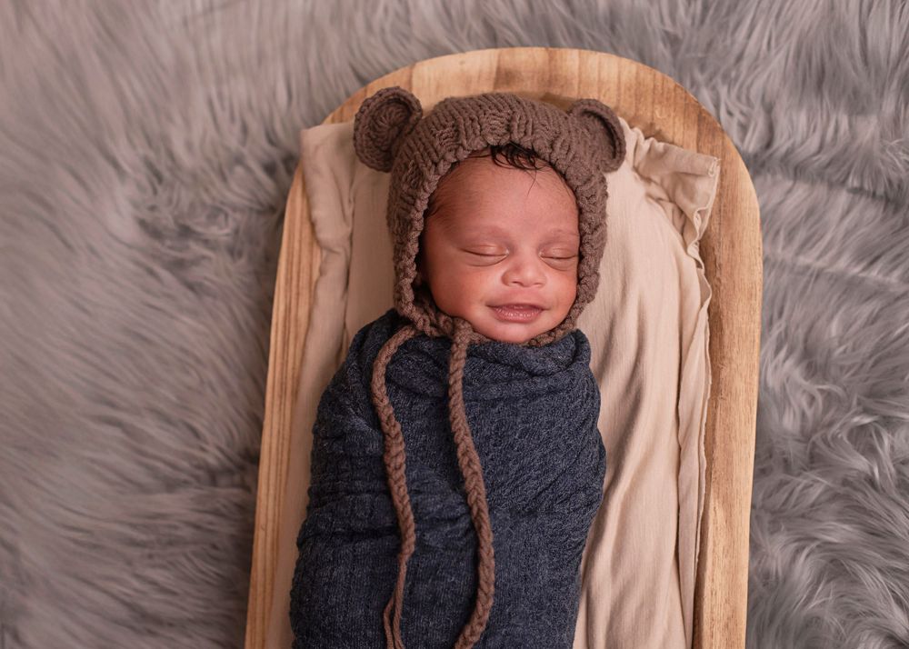 Smiling newborn baby swaddled in blue knit wrap wearing brown bear bonnet, sleeping in wooden bed on gray fur.