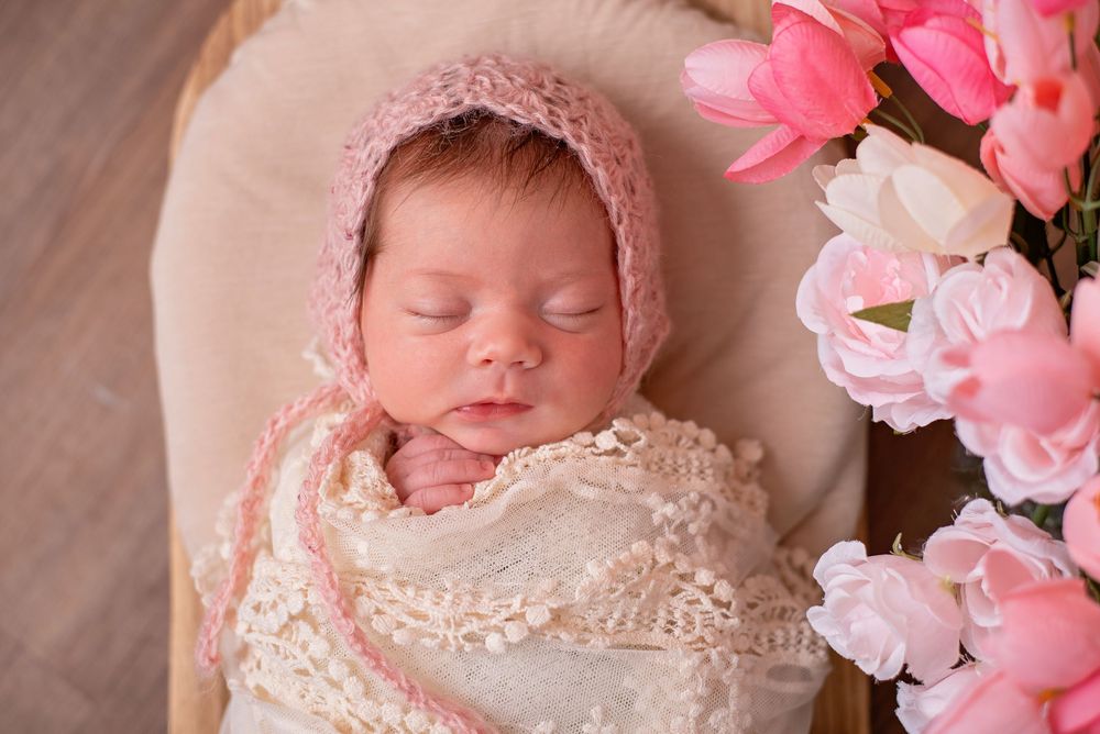 Sleeping newborn in cream lace dress surrounded by delicate pink flowers and soft lighting.