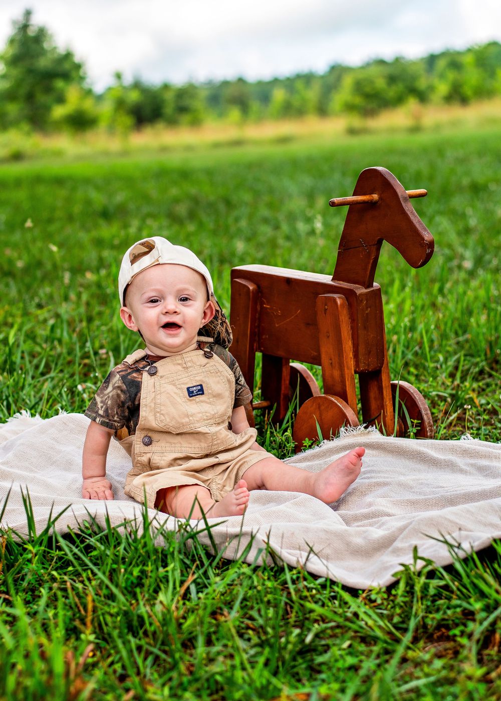 A smiling baby in tan overalls and a cap sits on a blanket in a green field beside a wooden toy horse.