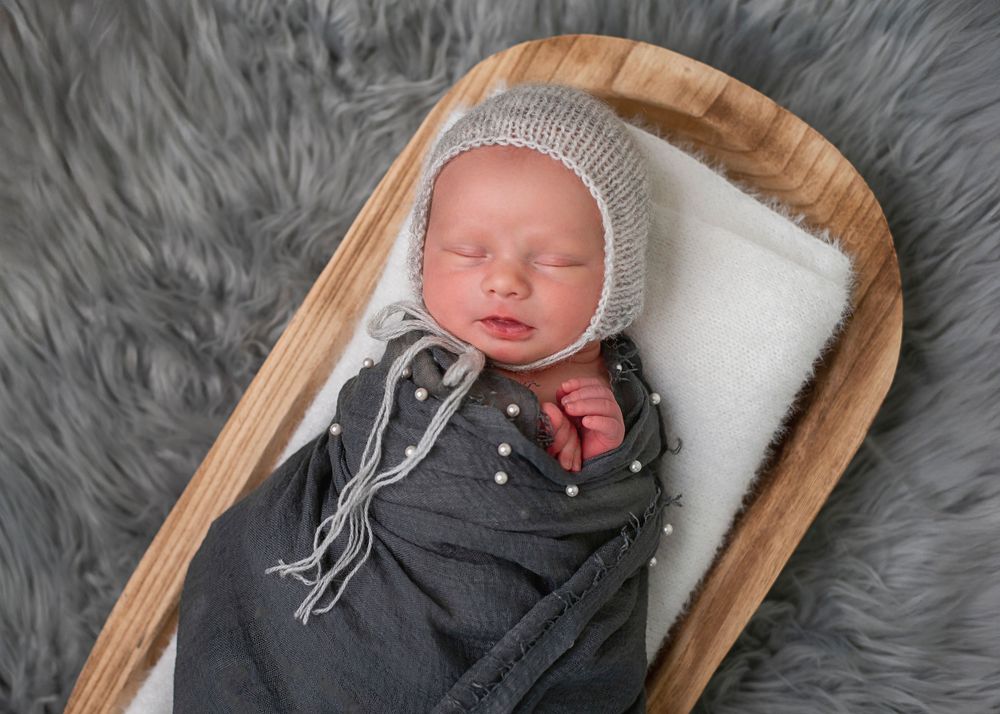 Sleeping newborn baby wrapped in gray swaddle, wearing knit bonnet, resting in wooden bowl on fluffy gray rug.