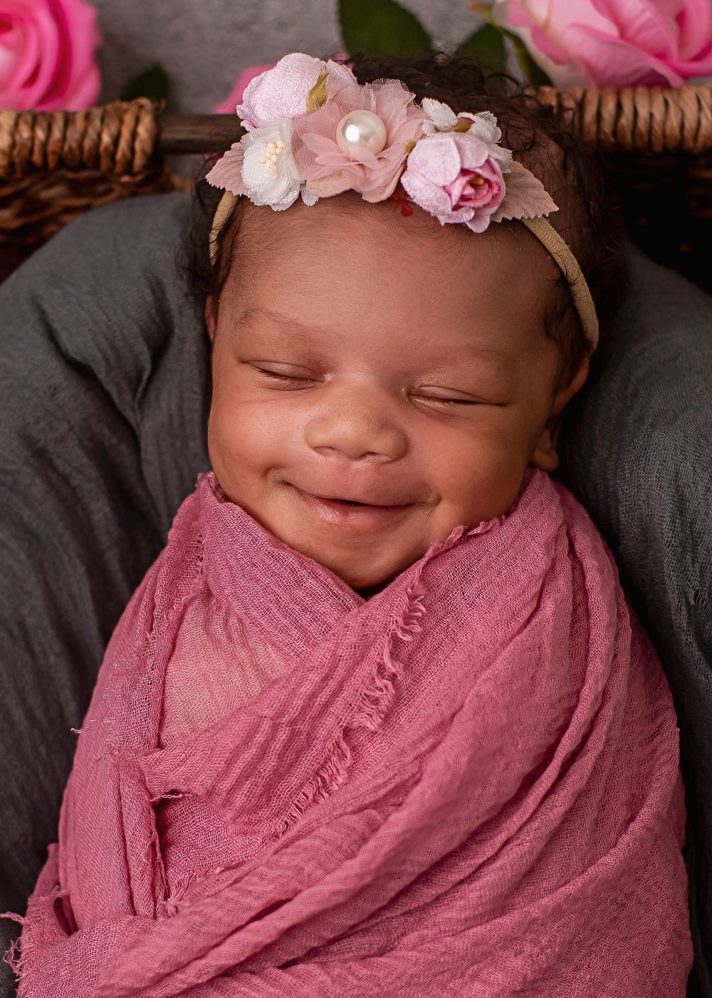 Smiling newborn baby girl swaddled in pink wrap wearing floral headband, sleeping peacefully in basket.
