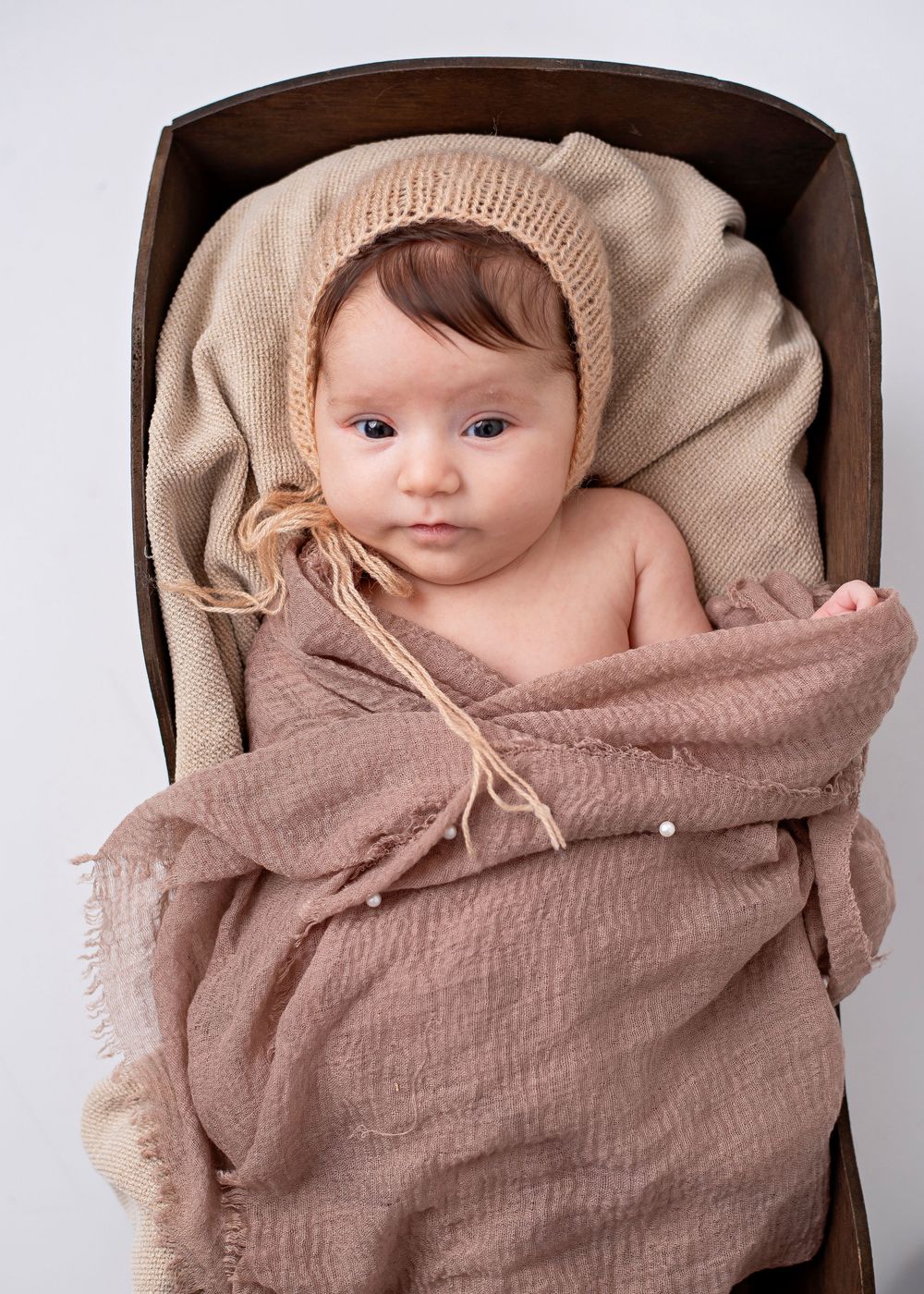 A baby wearing a knit beanie lies swaddled in soft beige and brown textured fabric against a wooden backdrop.