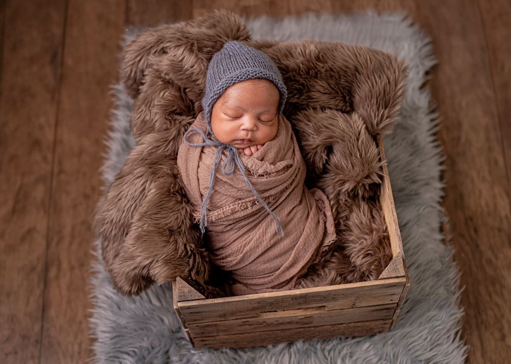 Newborn baby swaddled in brown wrap and blue bonnet sleeping in a wooden crate on fluffy fur rug.
