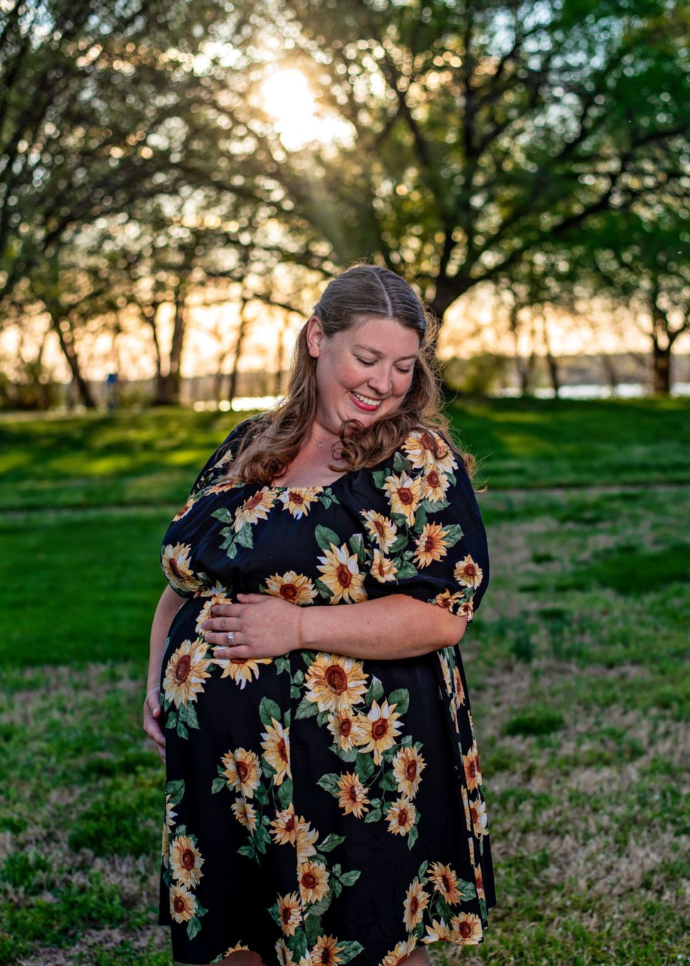 Pregnant woman in black sunflower dress cradling baby bump at golden hour in a lush green park.