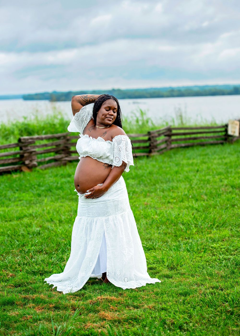 Pregnant woman in white lace off-shoulder dress cradling baby bump outdoors near a wooden fence and lake.