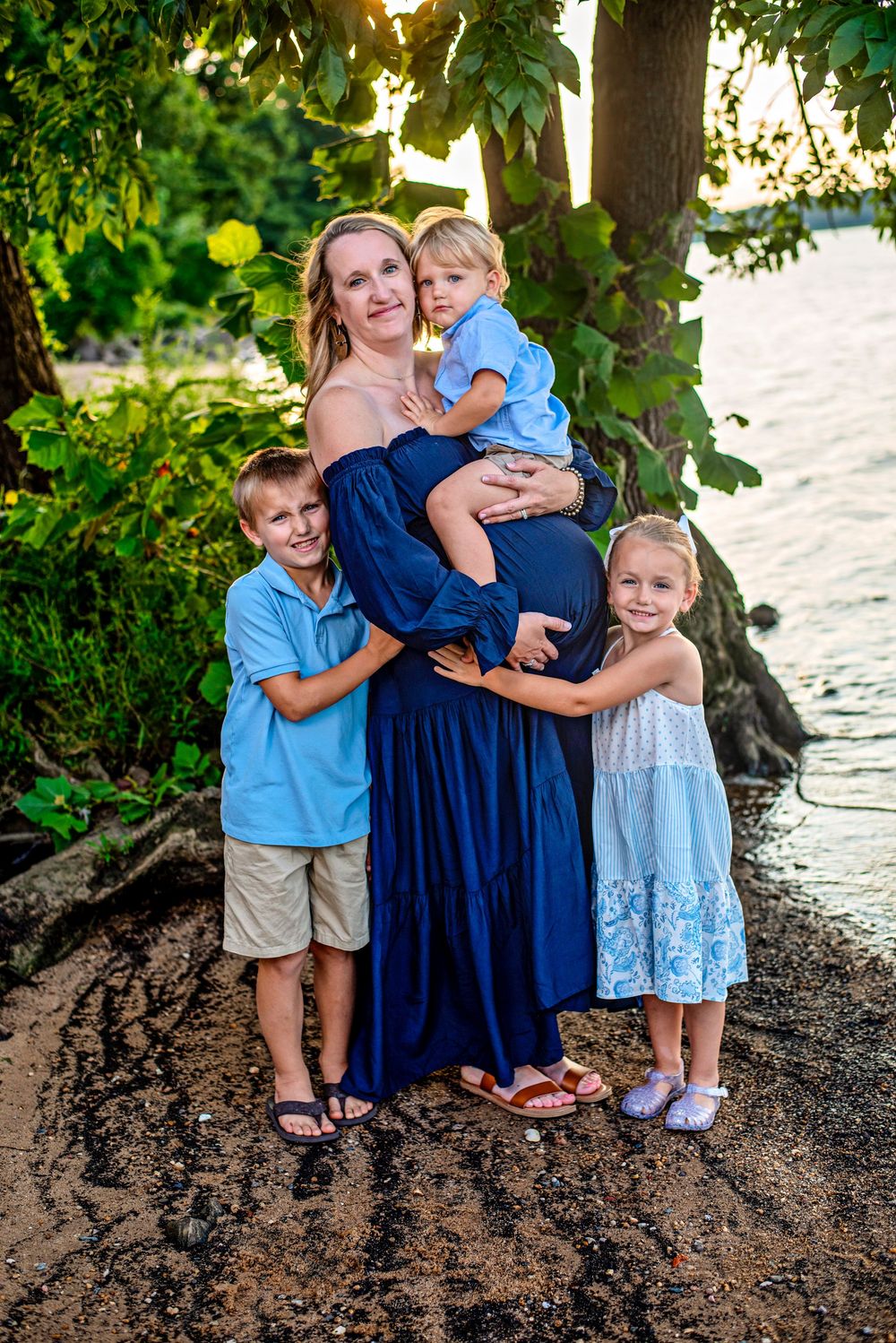 Pregnant woman in navy dress poses with three young children near tropical waterfront at golden hour.