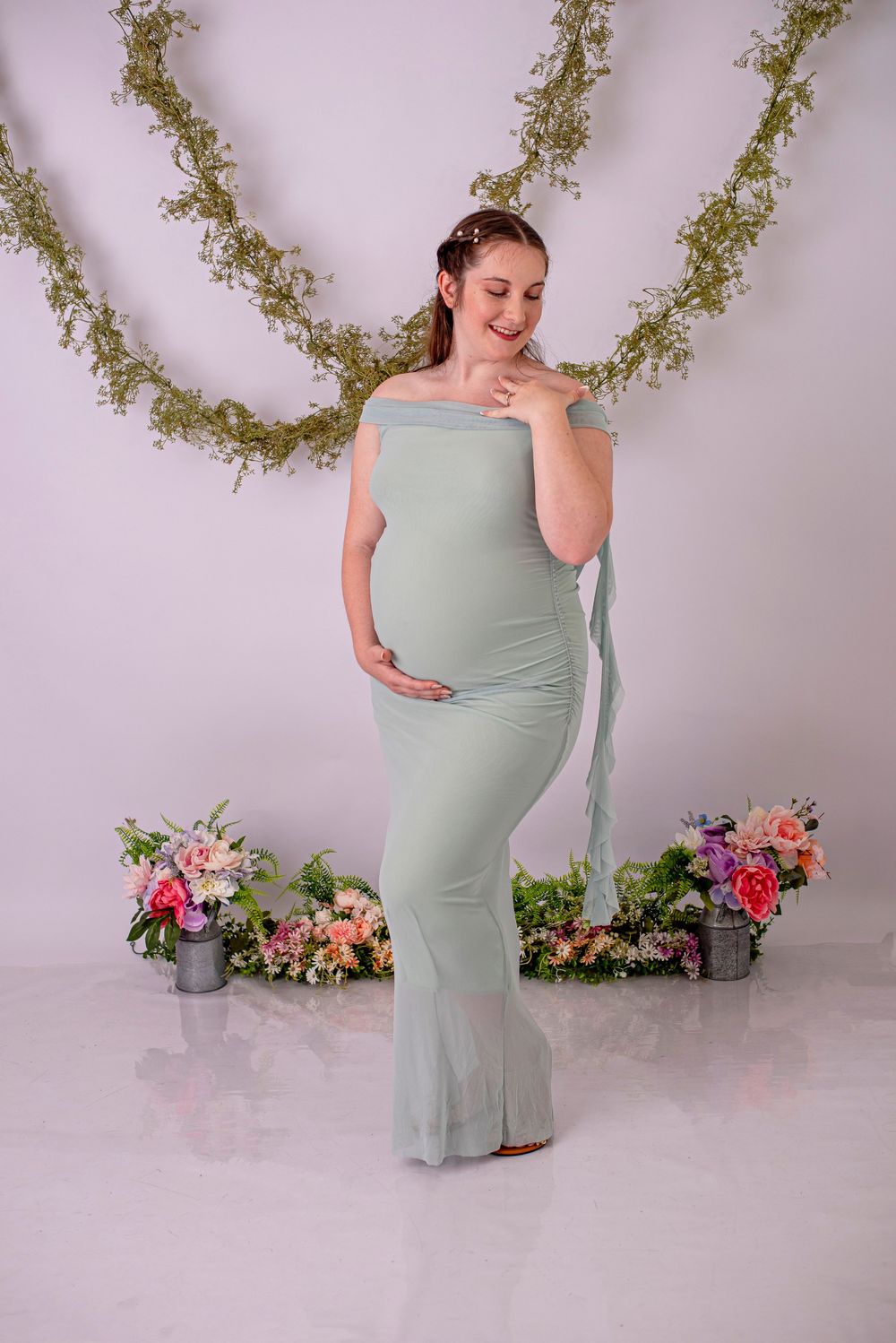 Pregnant woman in sage green off-shoulder maxi dress poses in studio with floral backdrop and greenery.