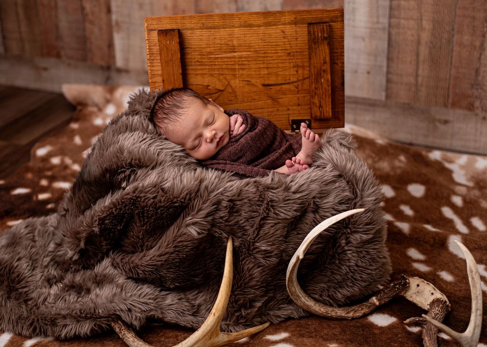 Newborn baby swaddled in brown wrap sleeping peacefully on fur blanket in rustic wooden crate with antlers.