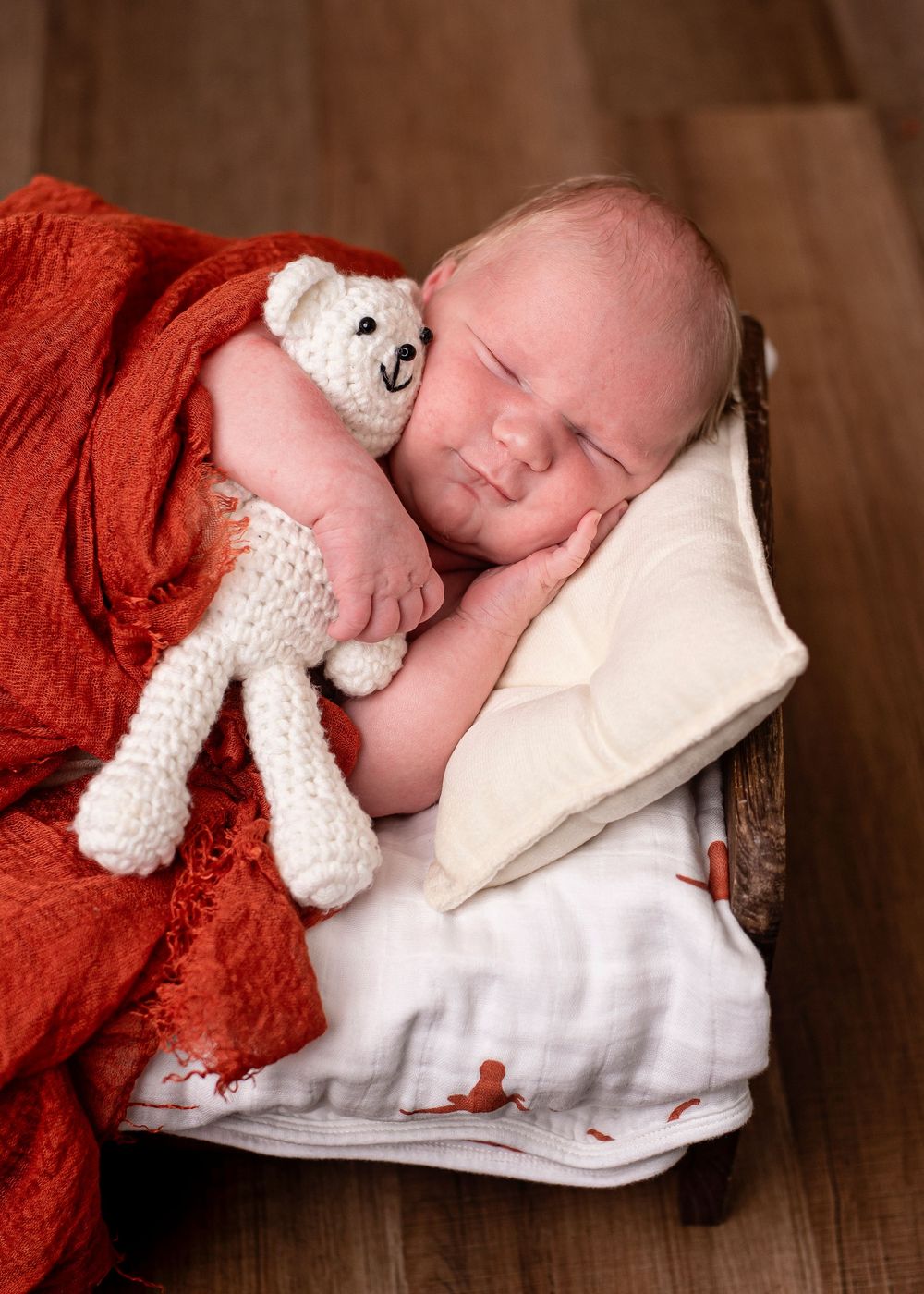 Sleeping newborn baby cuddling a white knitted teddy bear on a tiny bed with red blanket.