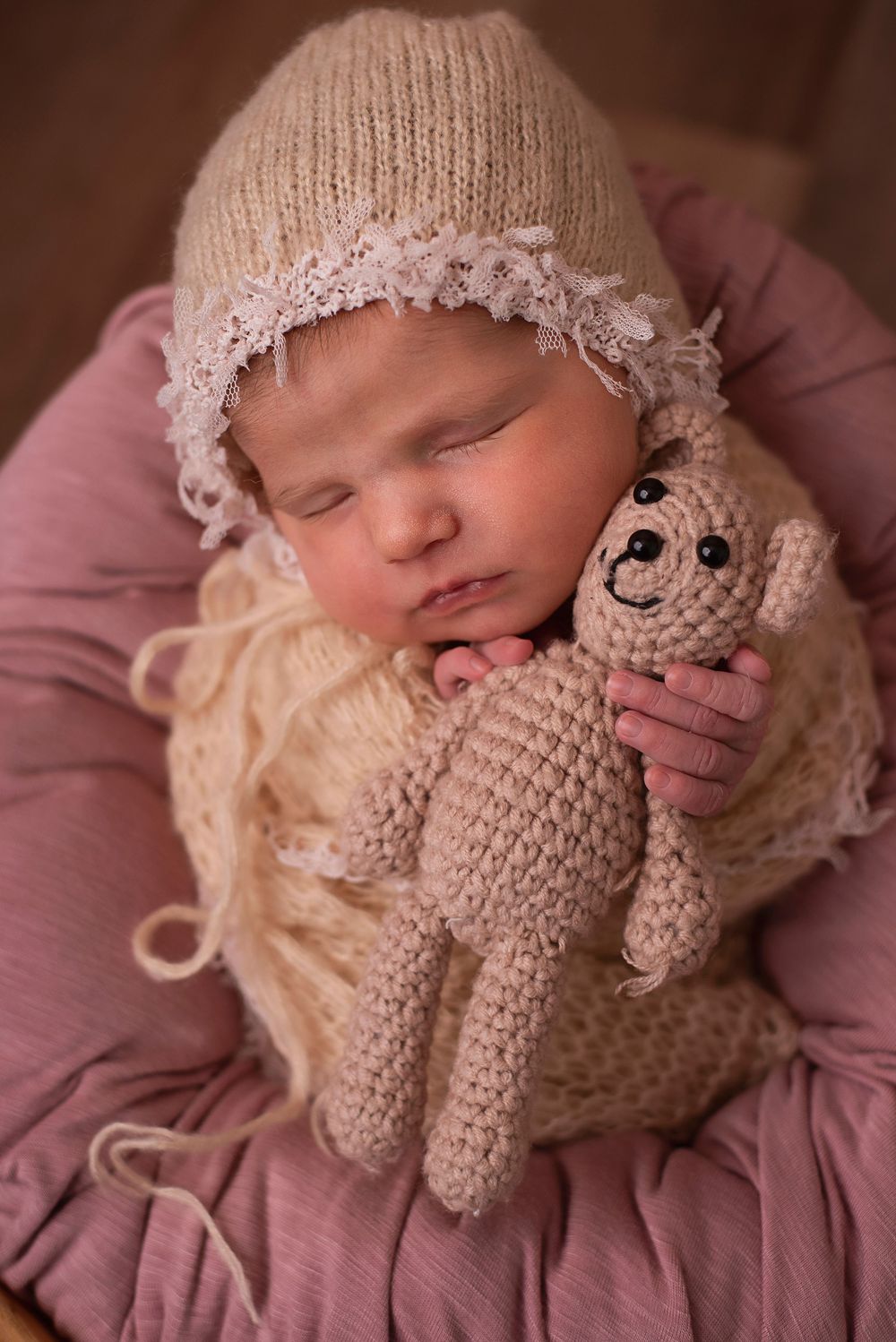 Sleeping newborn baby in knit bonnet cuddling a crocheted teddy bear on a pink cushion.