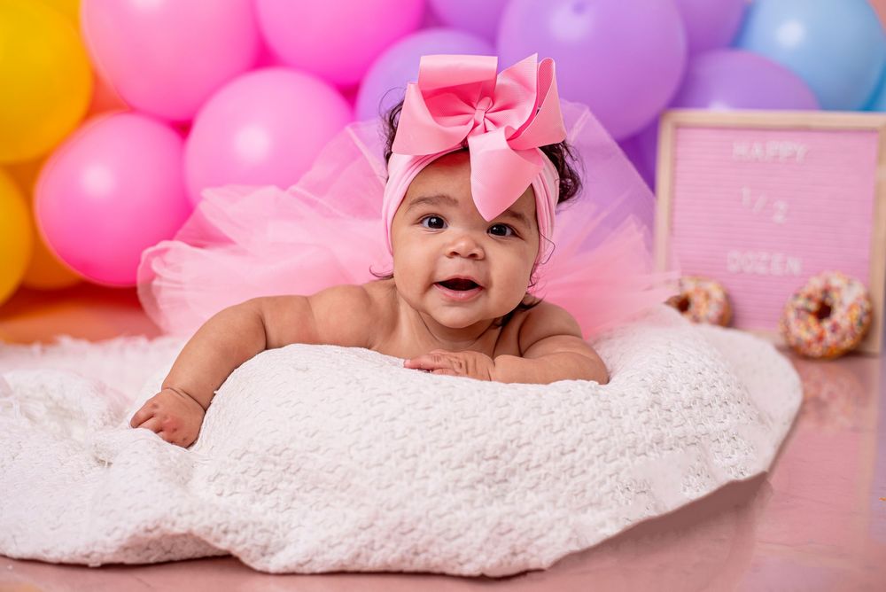 Baby girl in pink tutu headband smiles on white blanket surrounded by colorful balloons at her first birthday.