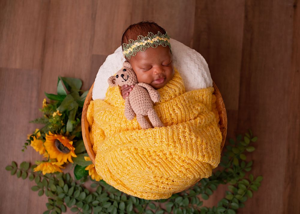 A sleeping newborn nestled in a bright yellow knitted blanket with sunflowers and greenery as decorative accents.