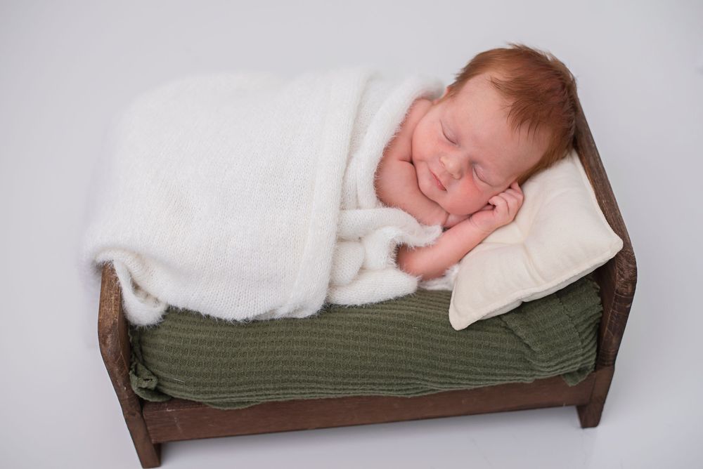 Sleeping newborn baby wrapped in white blanket on tiny wooden bed with green mattress and cream pillow.