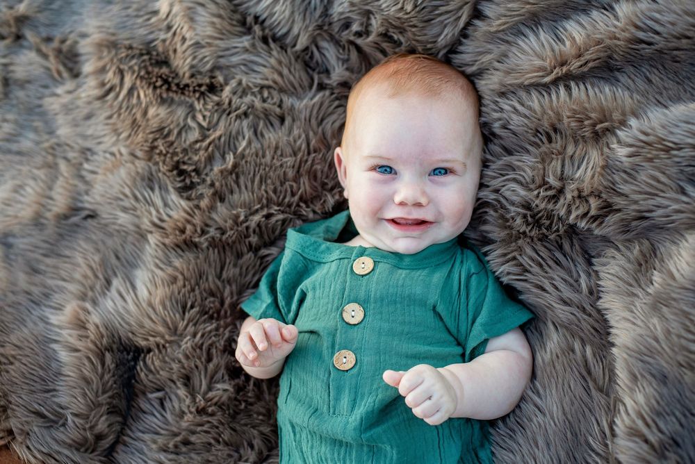 Baby wearing green button-up romper lies on soft gray faux fur blanket while smiling.