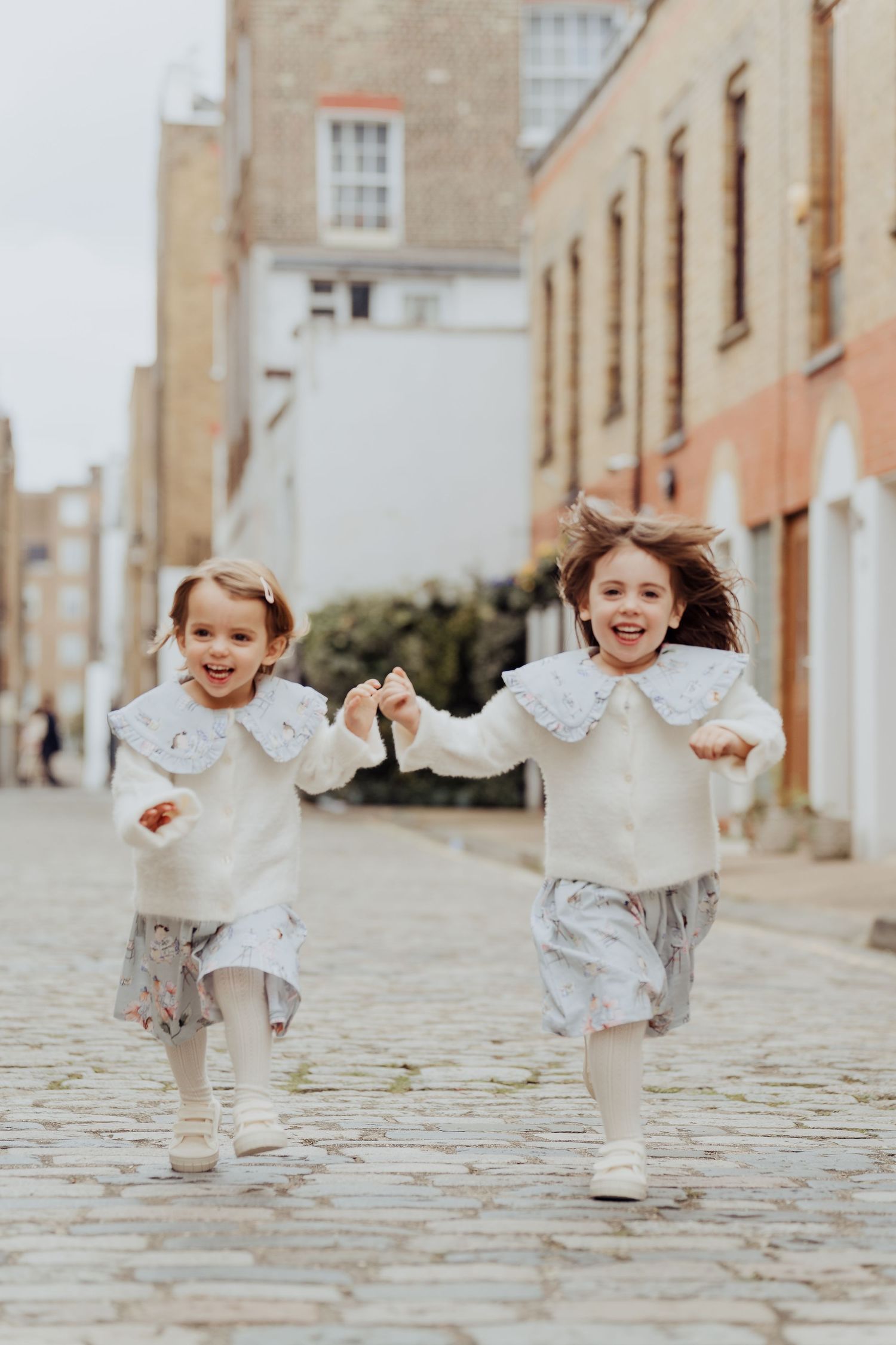 Two young children in white outfits run and play together on a cobblestone street lined with historic buildings.