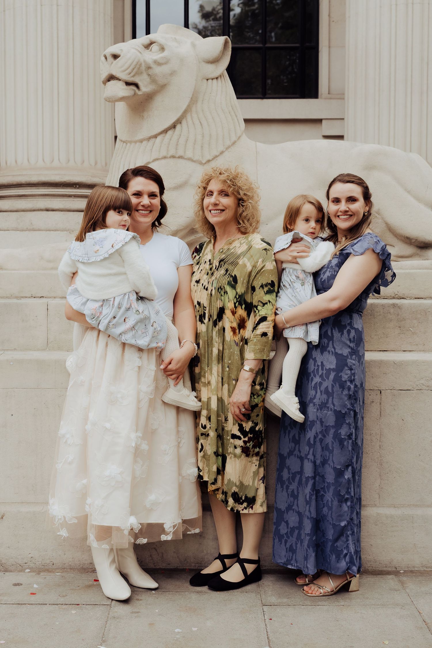 Three young children in vintage-style dresses pose together against a light-colored stone wall.