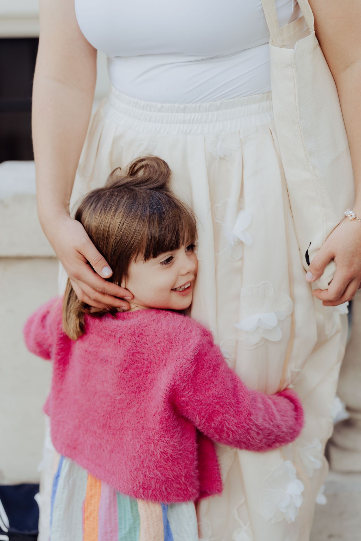 A small child wearing a pink fuzzy sweater and rainbow skirt hugs someone wearing white pants.