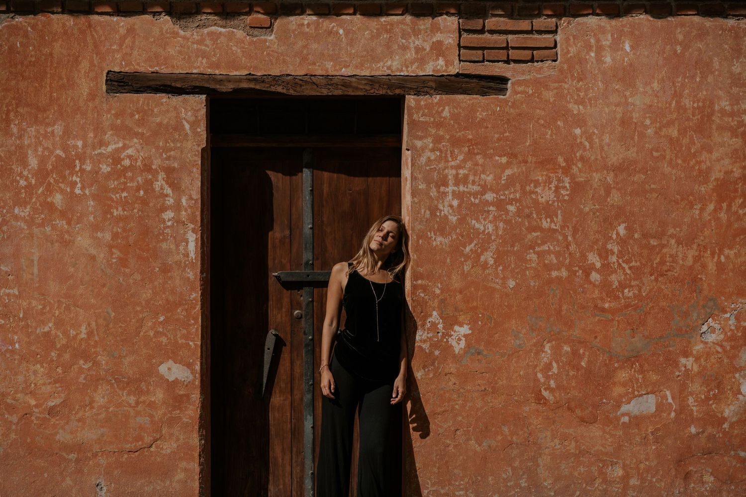 Person in a flowing red dress stands against a vibrant orange wall with hanging greenery.