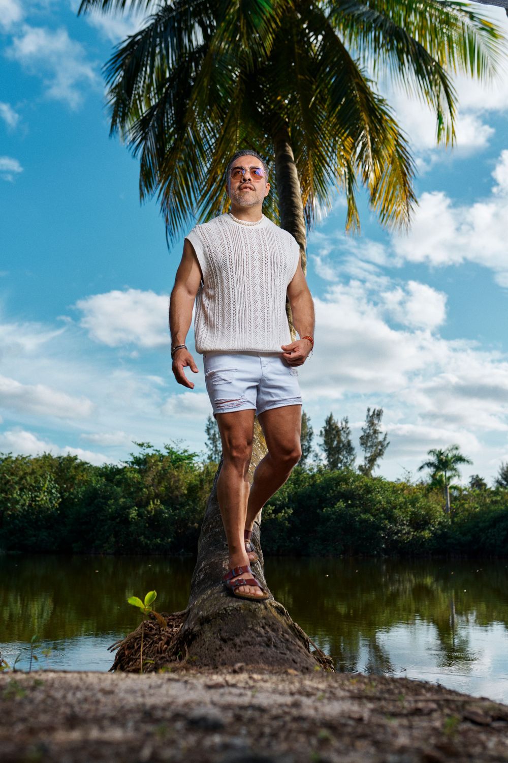 Athletic person in white shirt and shorts poses confidently on palm tree-lined riverbank.